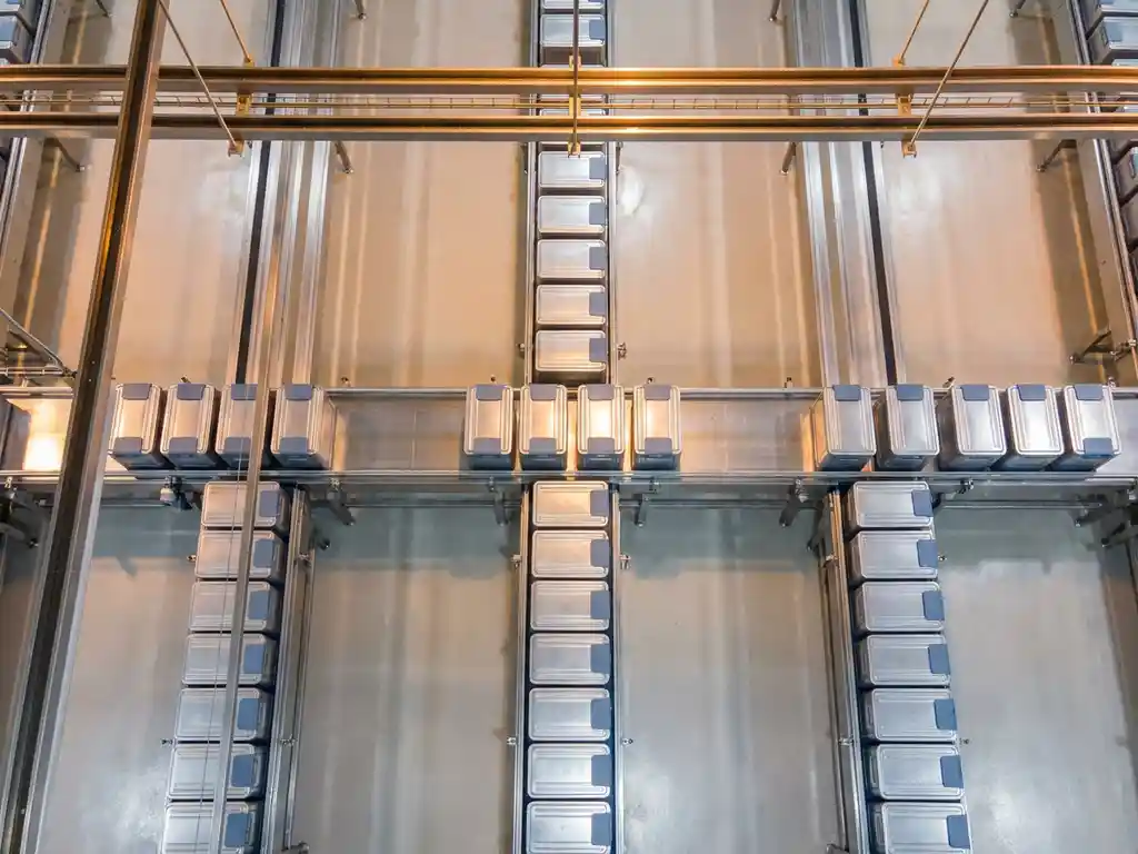 Identical industrial containers moving in uniform batches along a stainless steel conveyor belt in a clean factory, shot from overhead with warm amber lighting.